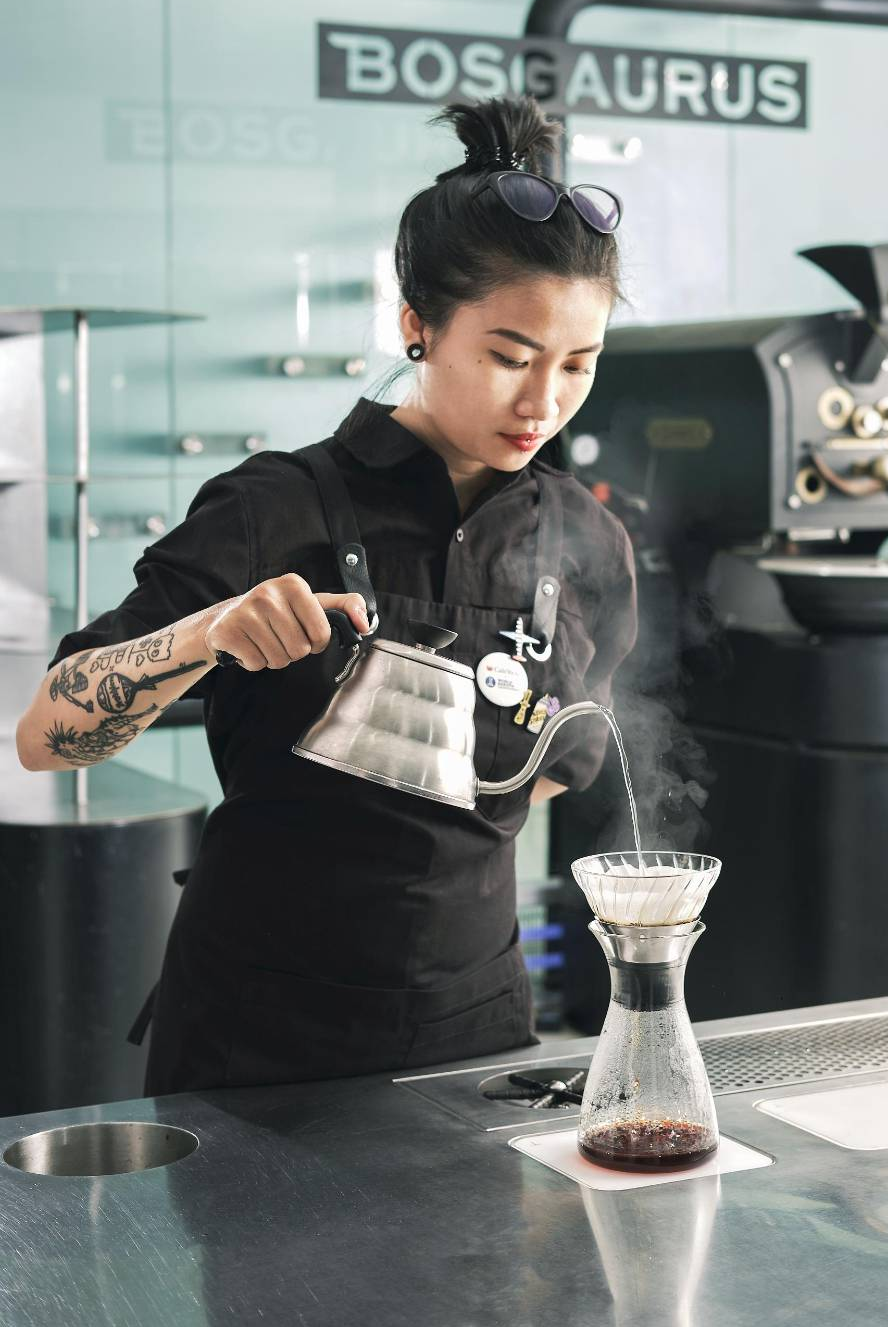 Barista with tattooed arm pouring hot water from a gooseneck kettle into a pour-over coffee maker.